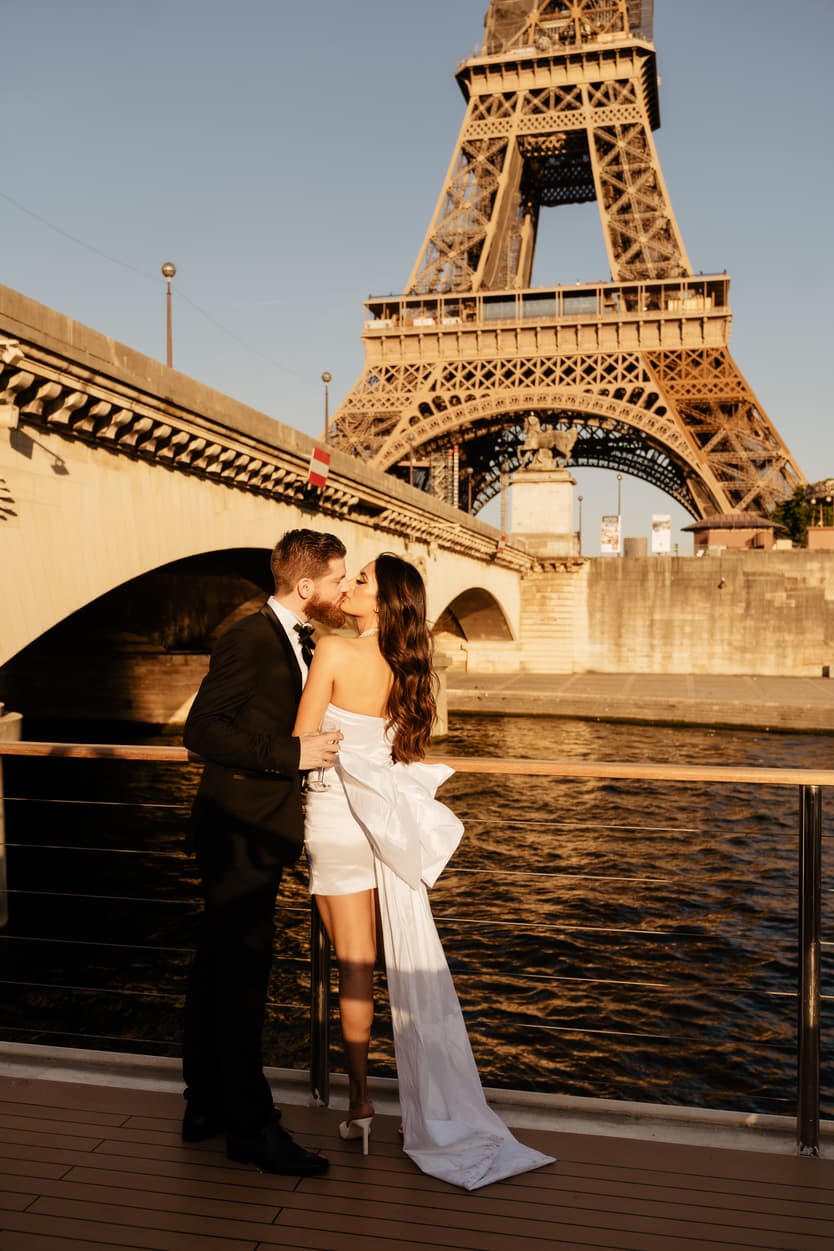 The couple kissing on the boat deck under the Eiffel Tower, her white welcome-dinner dress with its long bow