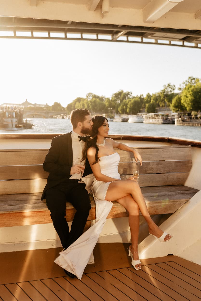 The couple sitting together on the boat’s bench, her white dress across the wooden deck