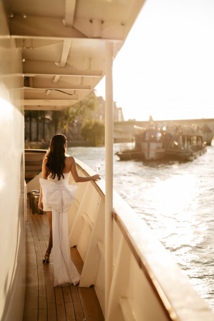 The bride walking the deck in a short white dress with a trailing bow, the Seine going by