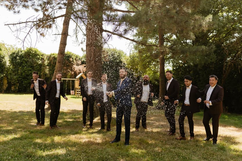The groom opening an oversized bottle of champagne on the grass, his groomsmen around him
