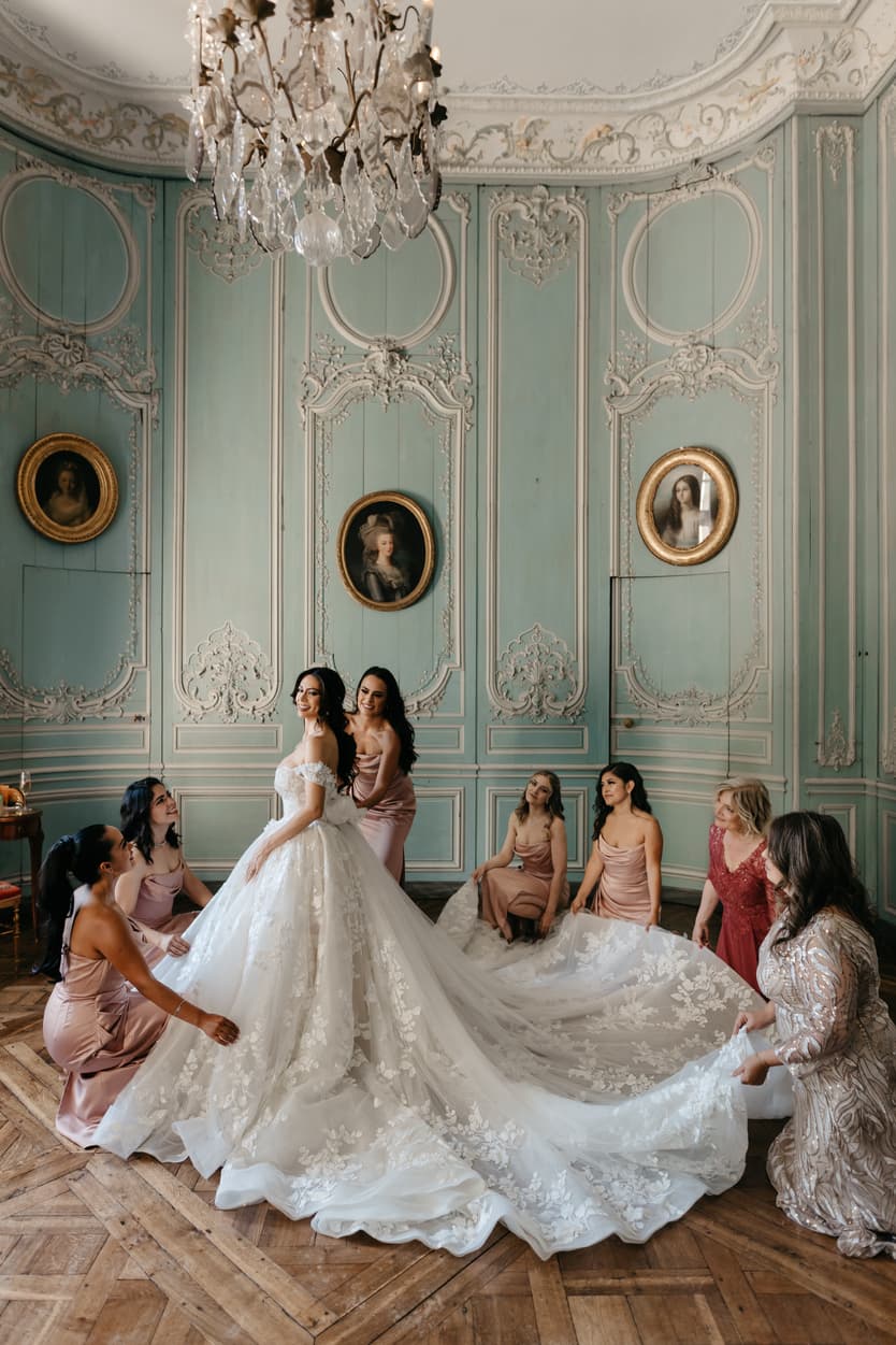The bride in her ivory lace ball gown with her bridesmaids in dusty-rose satin, laying out the train in the château’s green salon