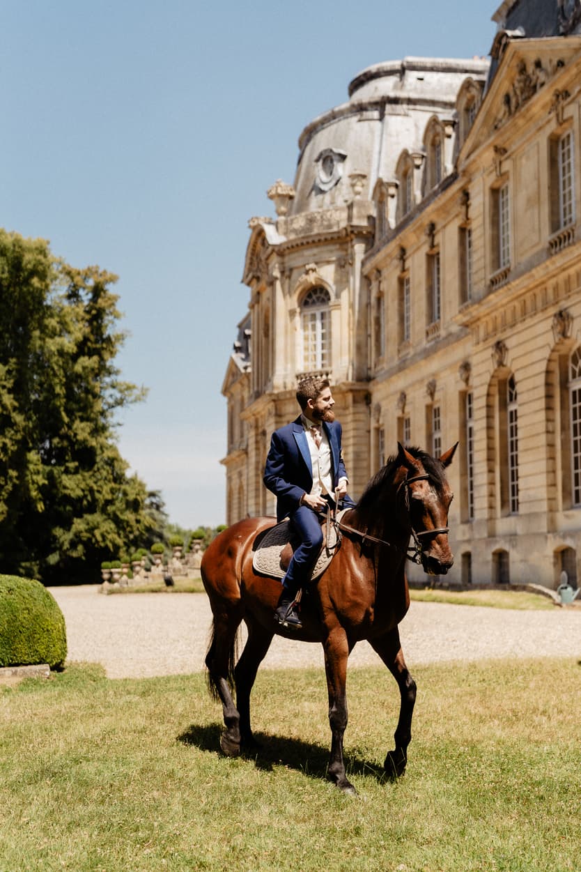 The groom riding a chestnut horse across the lawn in front of the château in his blue wedding suit