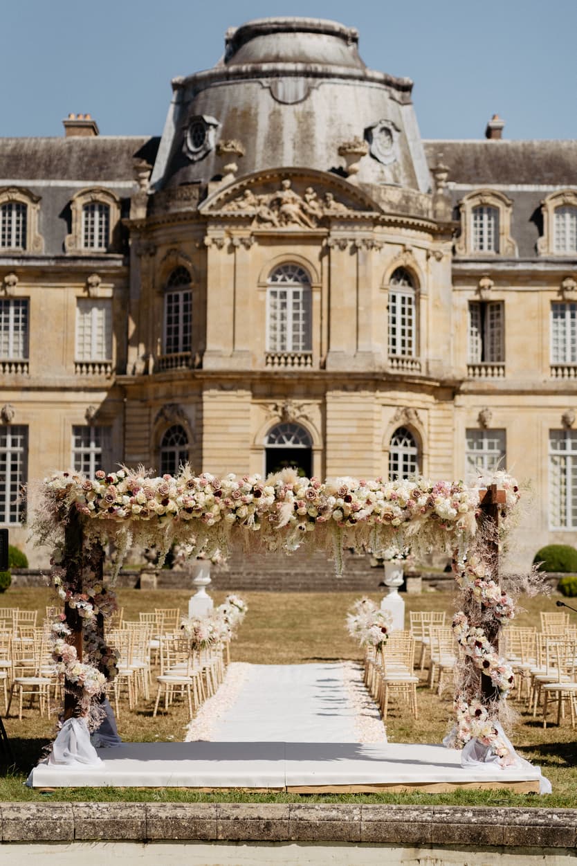 Wedding arch of blush and cream roses with pampas grass, the petal aisle and gold chairs before the château