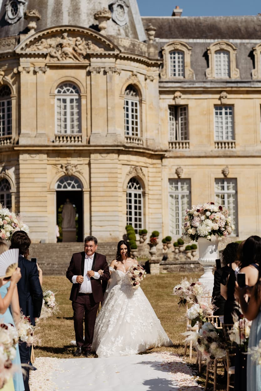 Her father walking the bride down the white aisle between the guests, the château ahead