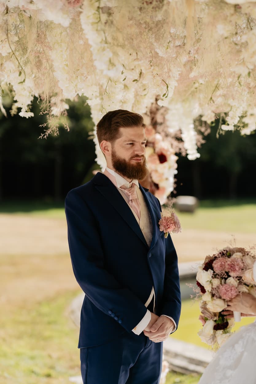 The groom under the flower arch watching the bride, the park of the château behind