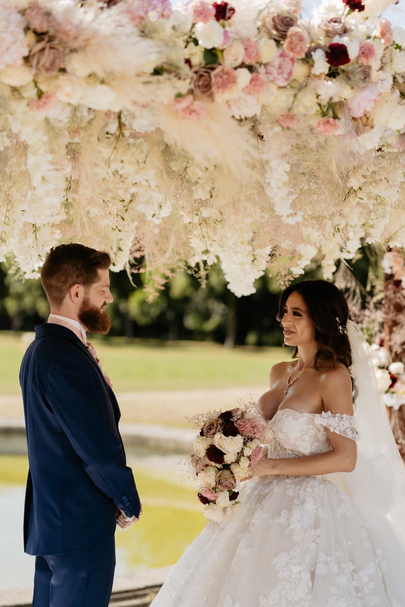 The couple at the arch of blush roses and pampas, her bouquet between them
