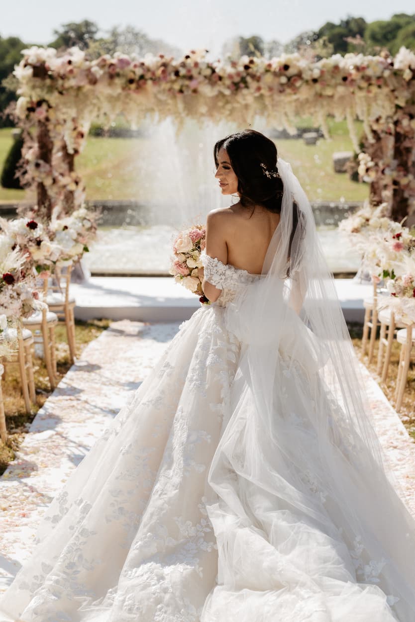 The bride from behind under the flower arch, the lace train and veil laid out on the white aisle