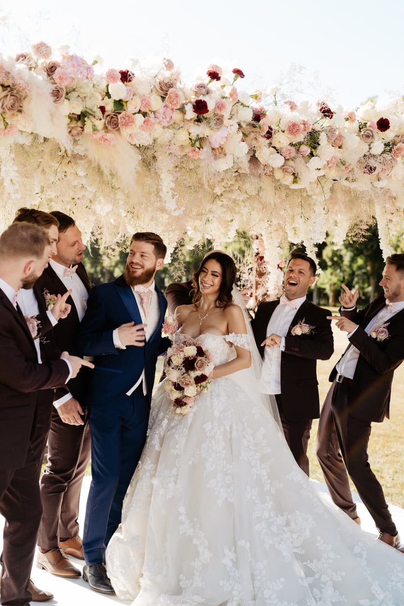 The wedding party around the couple under the flower arch, the groomsmen in black tie