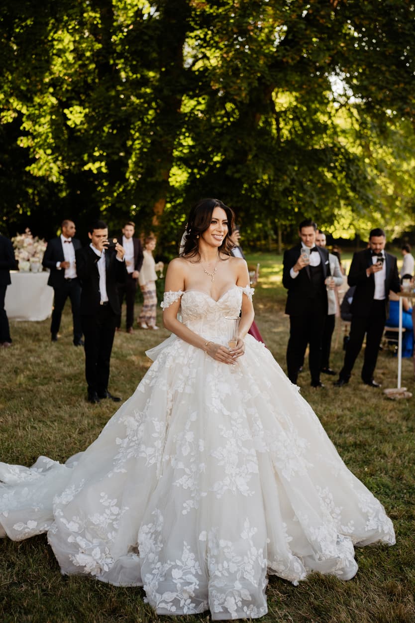 The bride alone on the grass in her lace ball gown, the guests standing behind her