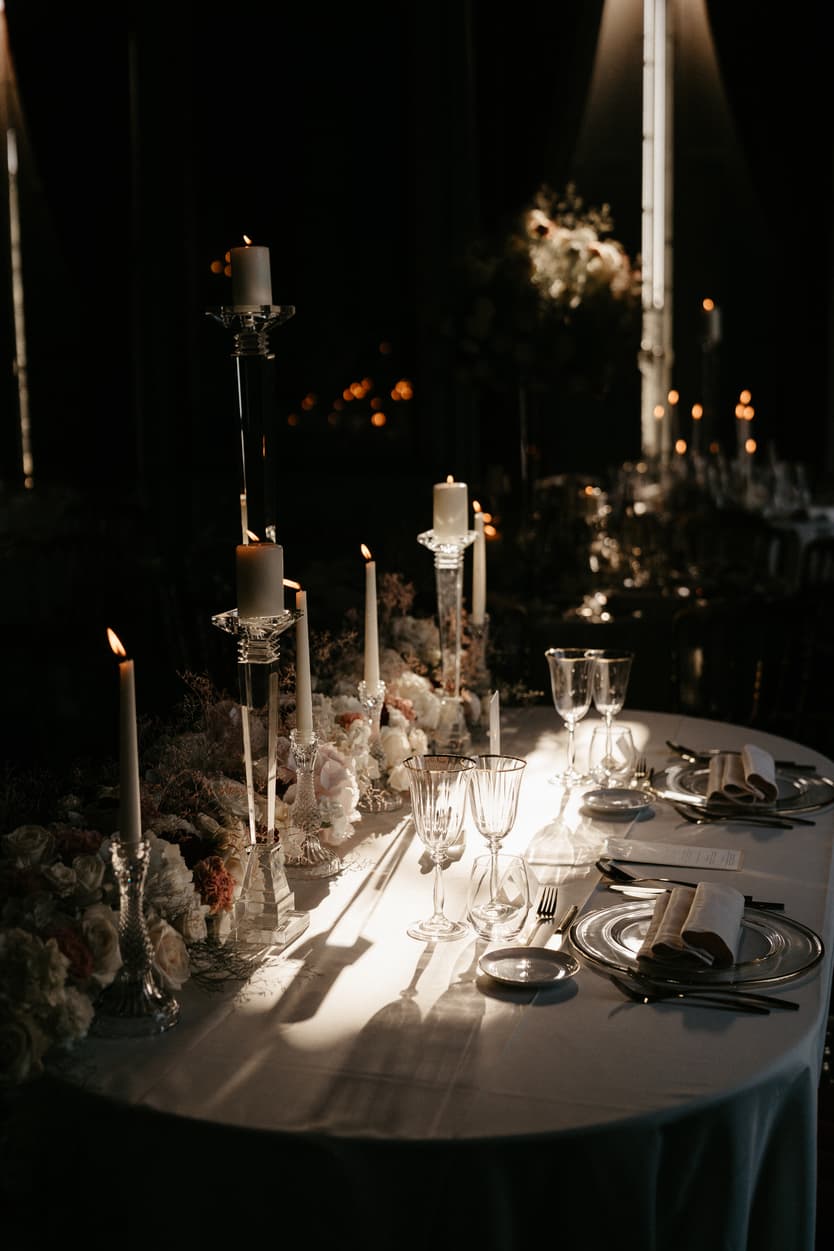 Tapered candles and glasses down the wedding table in the dark, the flowers lit from above