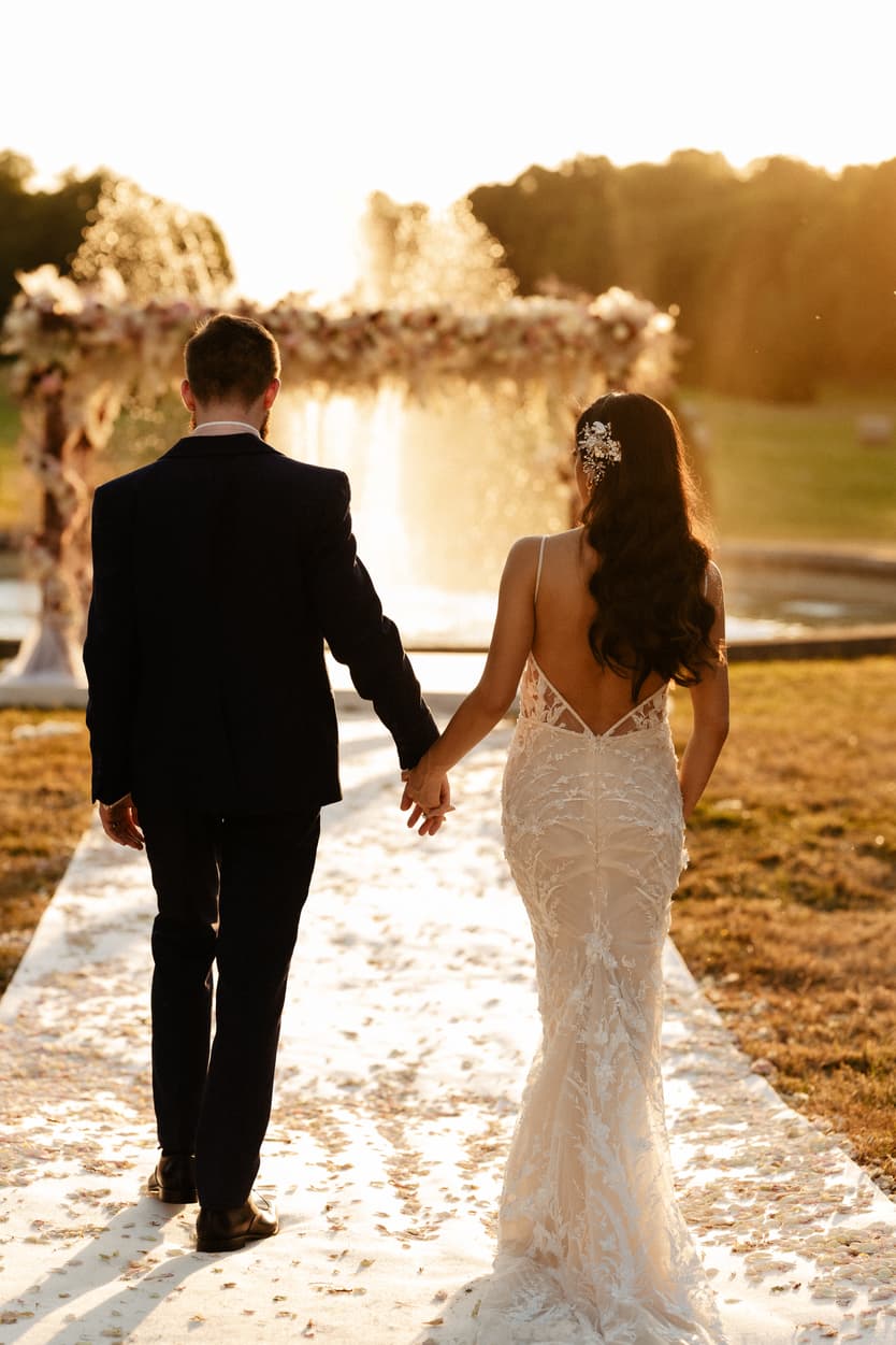 The couple walking the petal aisle towards the flower arch and the fountain at sunset