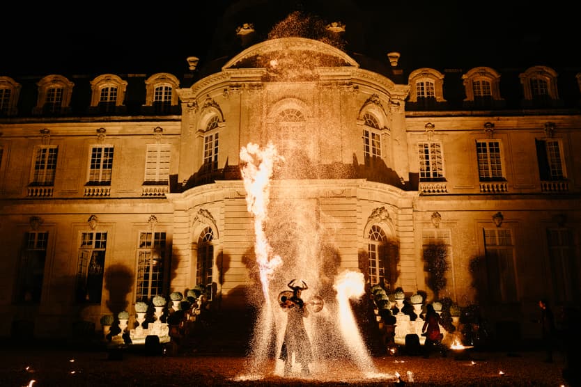 A fire performer spinning a burning frame before the lit façade of the château