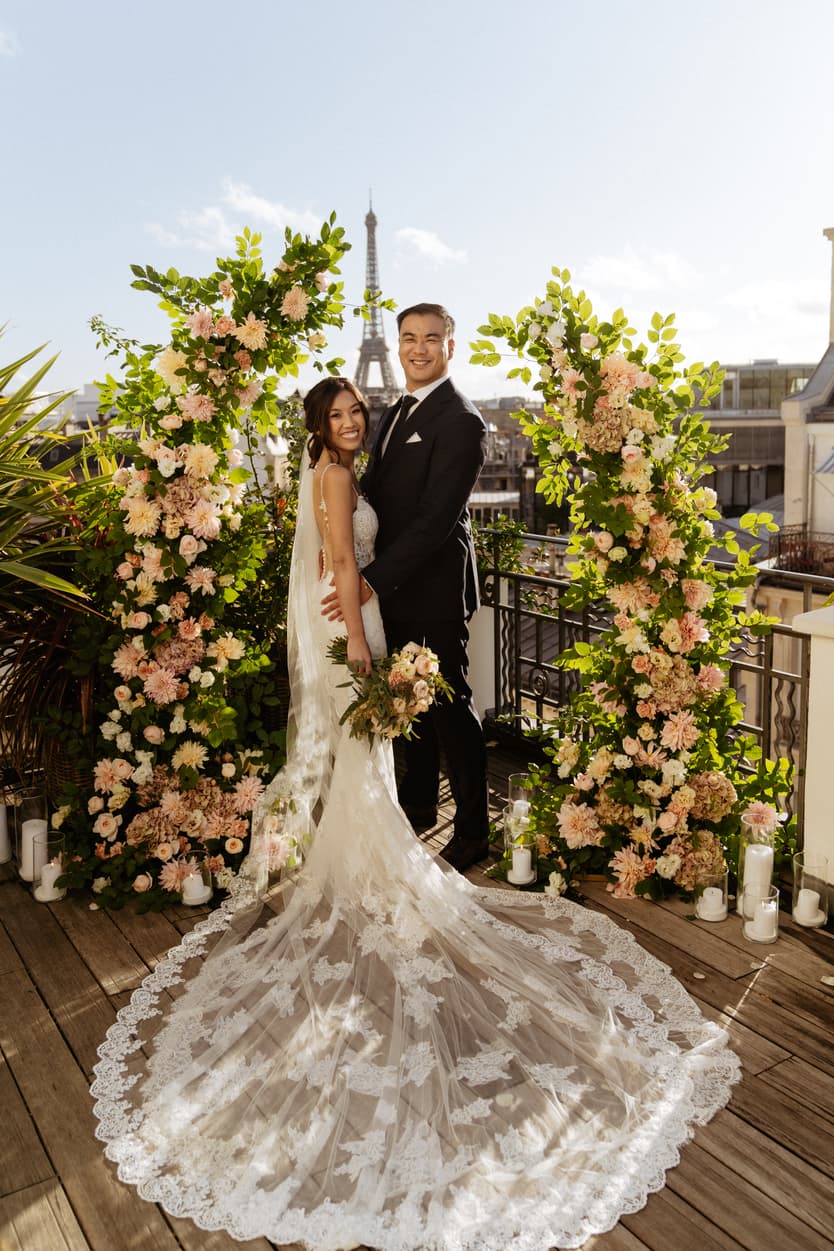 A couple under a flower arch on a Paris terrace, the bride’s embroidered train spread across the boards, the Eiffel Tower behind them