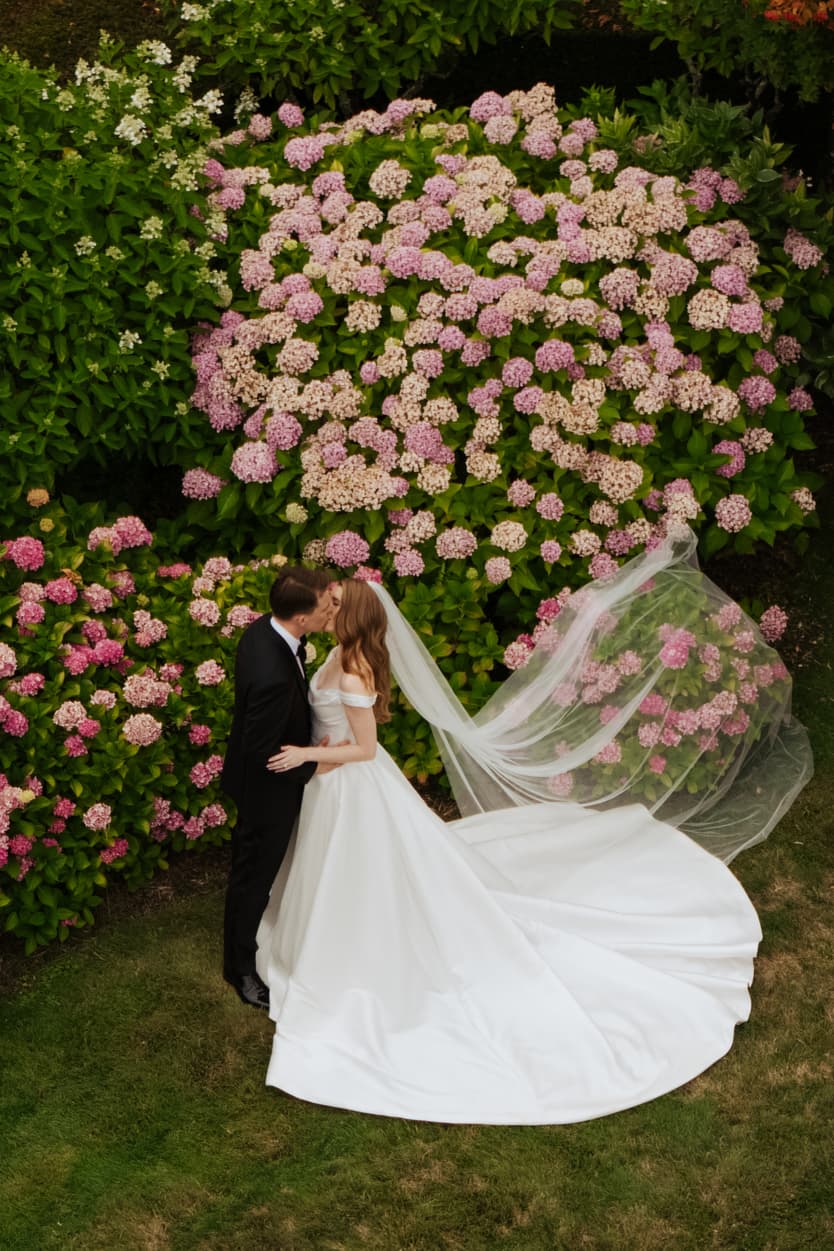 The couple kissing from straight above, her veil and satin train on the grass beside the pink hydrangea