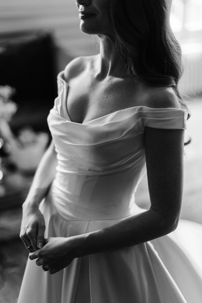 The bride’s off-shoulder satin wedding dress at a château window in black and white, close