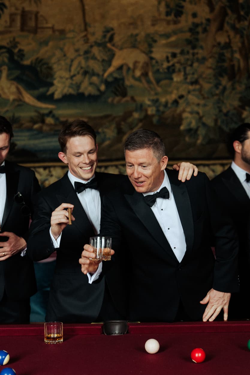 Groomsmen in black tie over a whisky at the billiard table, the tapestry of the château’s red room behind