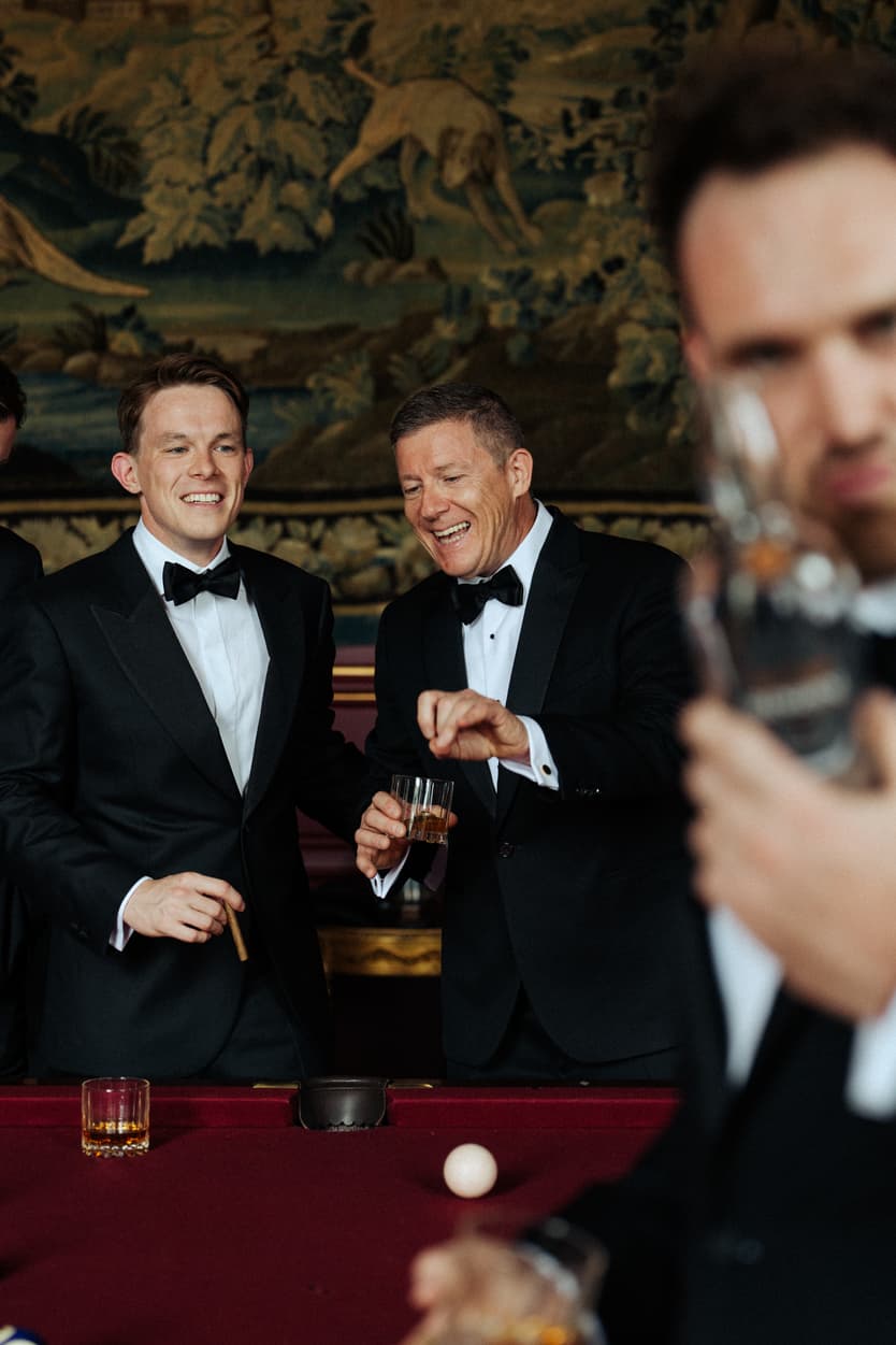 Two groomsmen laughing over a whisky at the billiard table before the château wedding