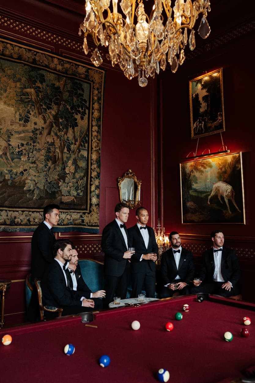 Groom and groomsmen in black tie around the billiard table at Château du Grand Lucé, under the chandelier