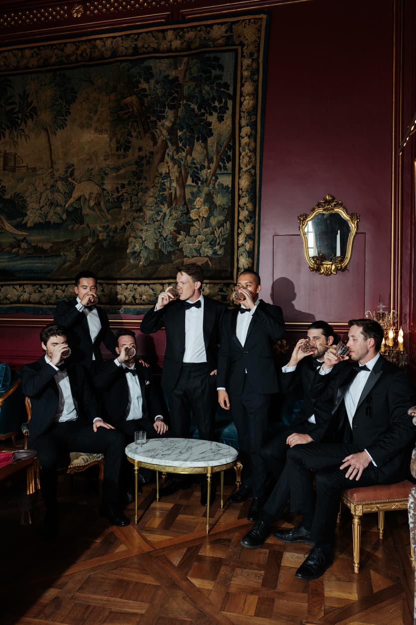 The groom and his groomsmen in black tie in the red room, tapestry and crystal chandelier above them