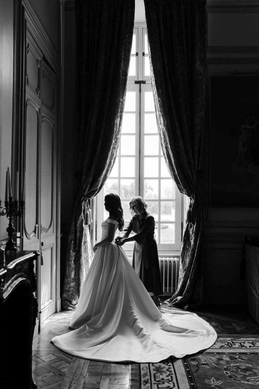 The bride and a bridesmaid at a tall window of Château du Grand Lucé in black and white, the train across the rug