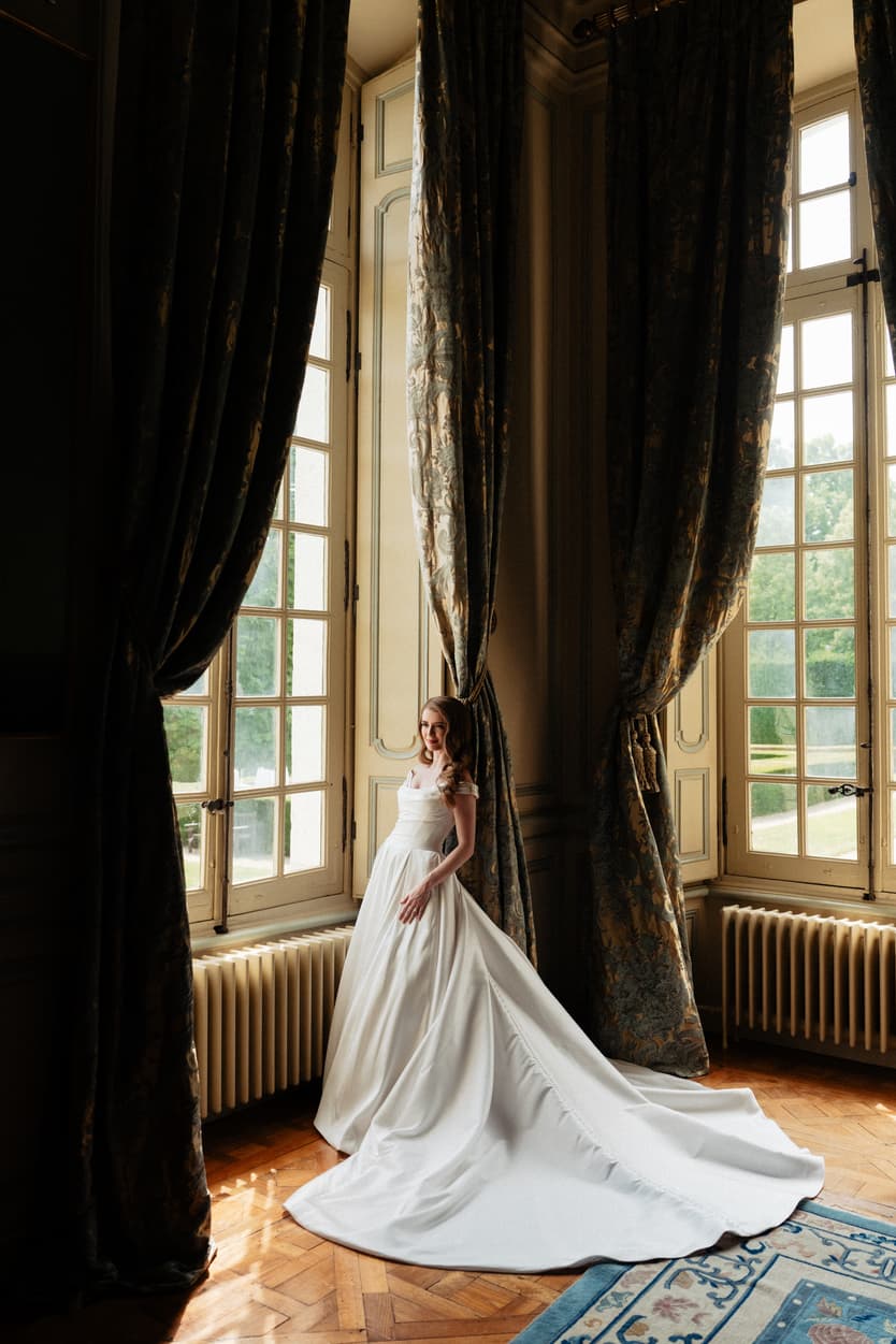 The bride in her ivory satin wedding dress at the château window, velvet curtains either side of her