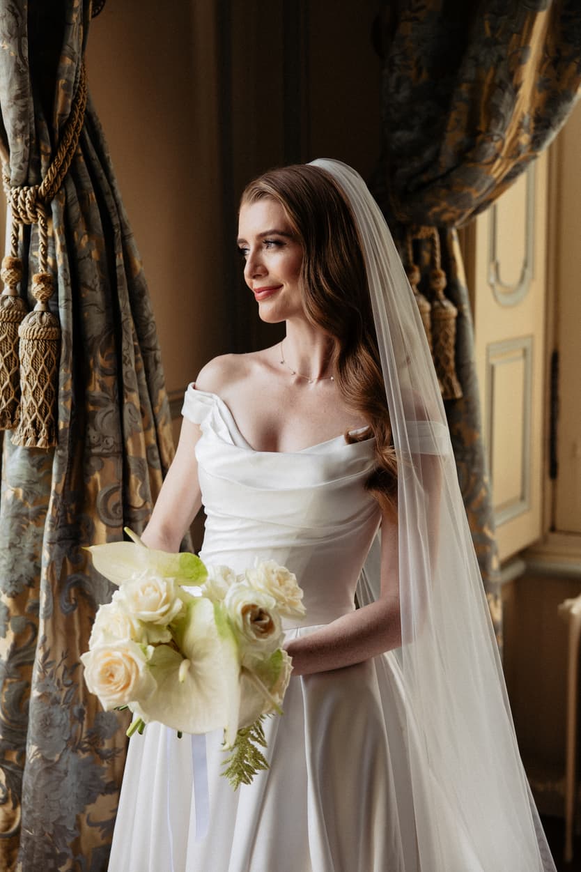 The bride in her off-shoulder satin wedding dress and veil at the château window with her white rose bouquet