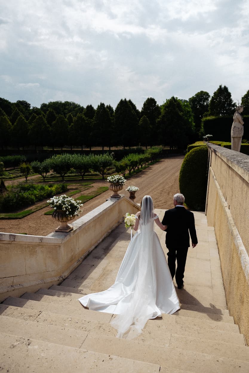 The bride and her father walking down to the ceremony at Château du Grand Lucé, her satin train on the steps