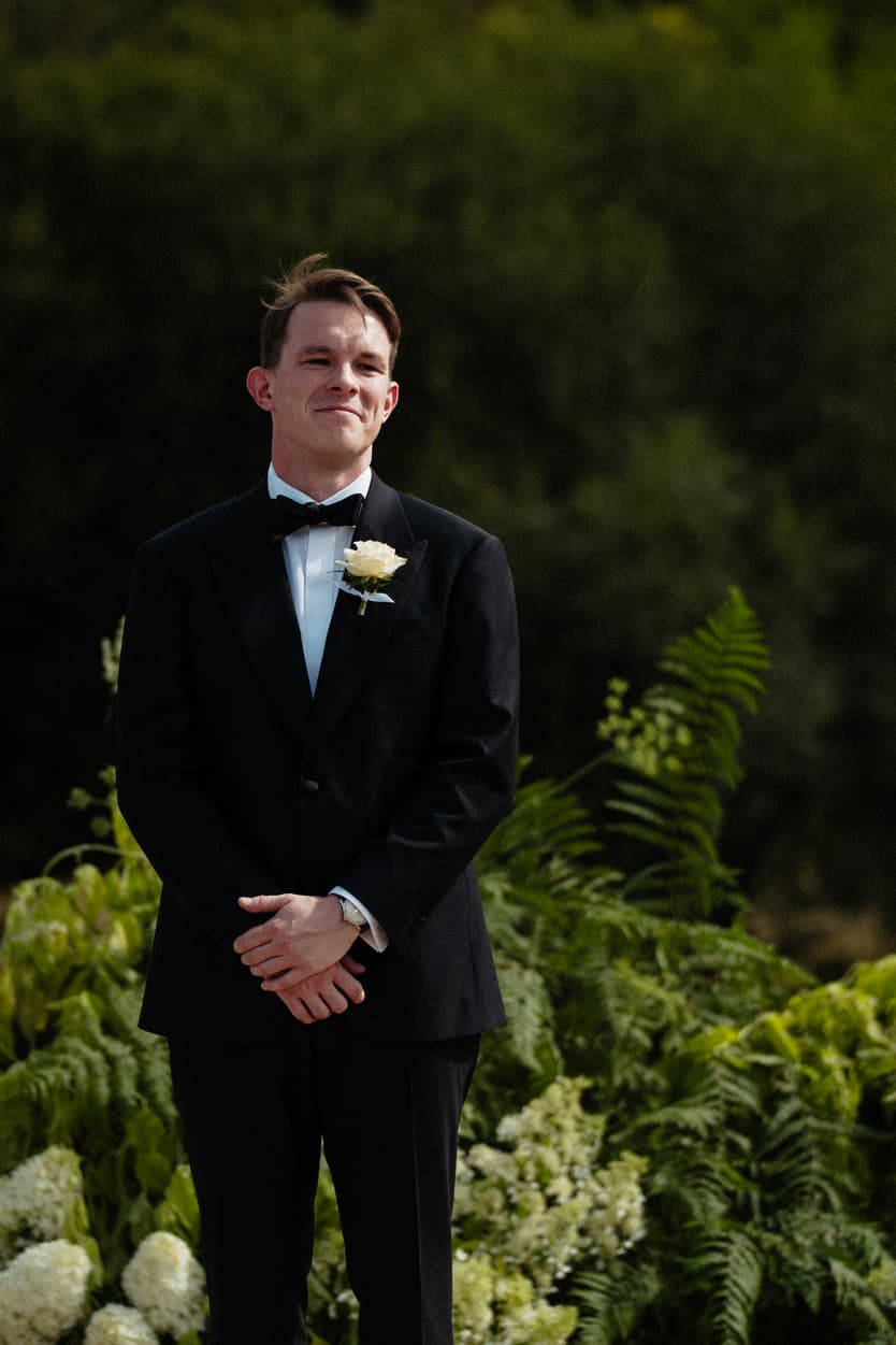 The groom at the end of the gravel aisle for the château ceremony, a white rose at his lapel and hydrangea behind