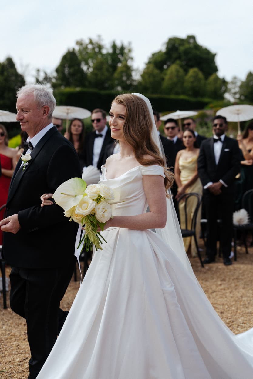 The bride on her father’s arm up the gravel aisle with her white bouquet, guests standing for the ceremony