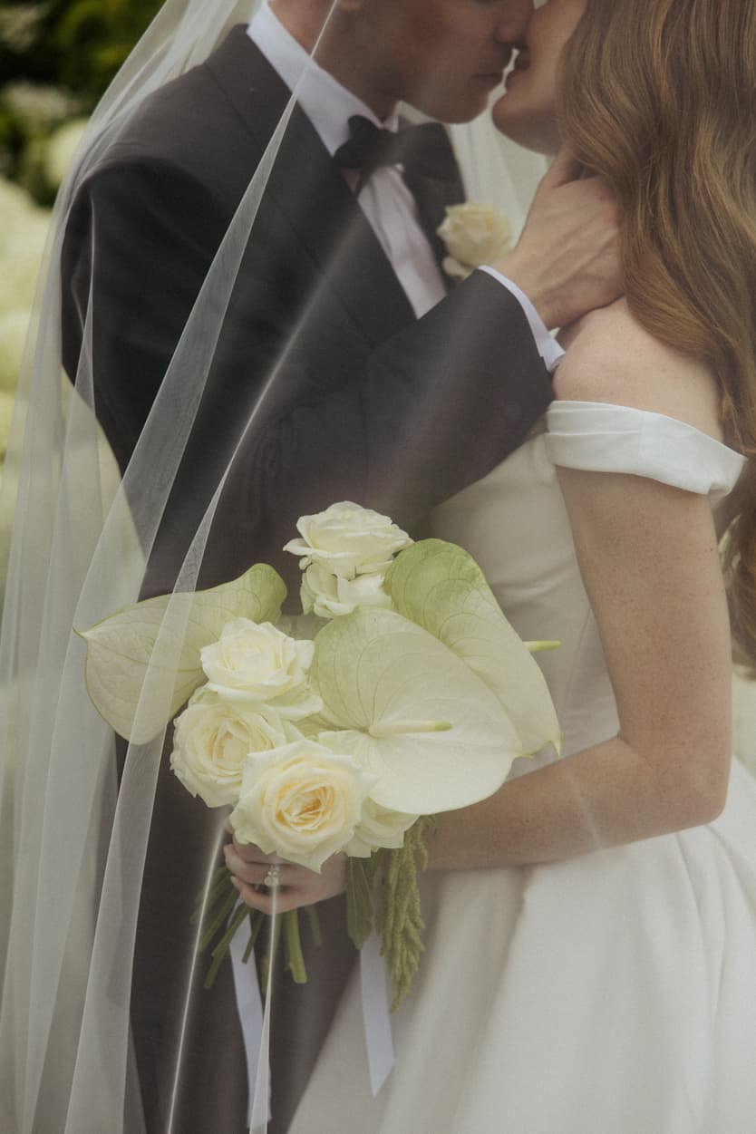 The bride’s white rose bouquet against her satin wedding dress under the veil, close