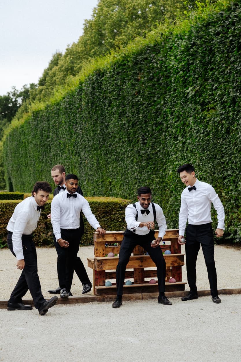 Groomsmen in bow ties and shirtsleeves playing a lawn game beside the tall topiary at Château du Grand Lucé