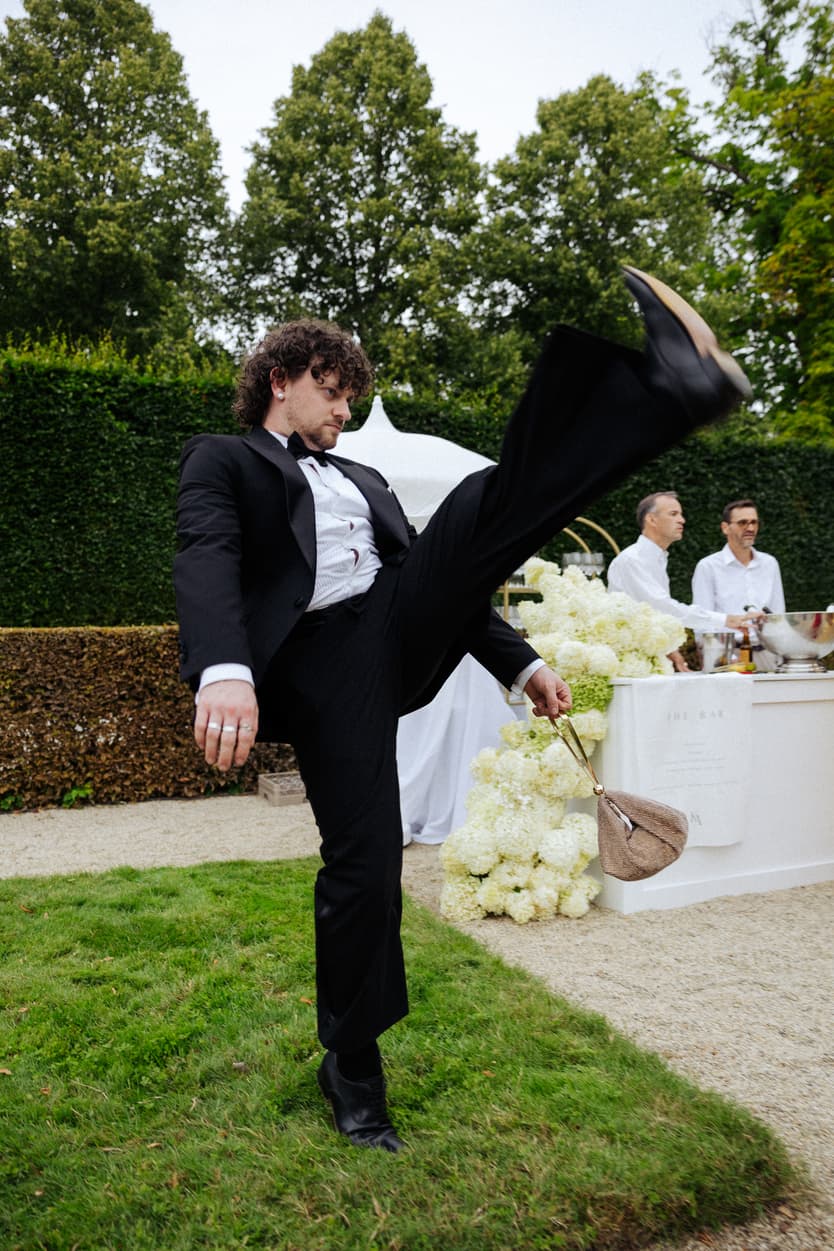A guest in black tie kicking a leg over his head on the lawn beside the bar banked with white hydrangea