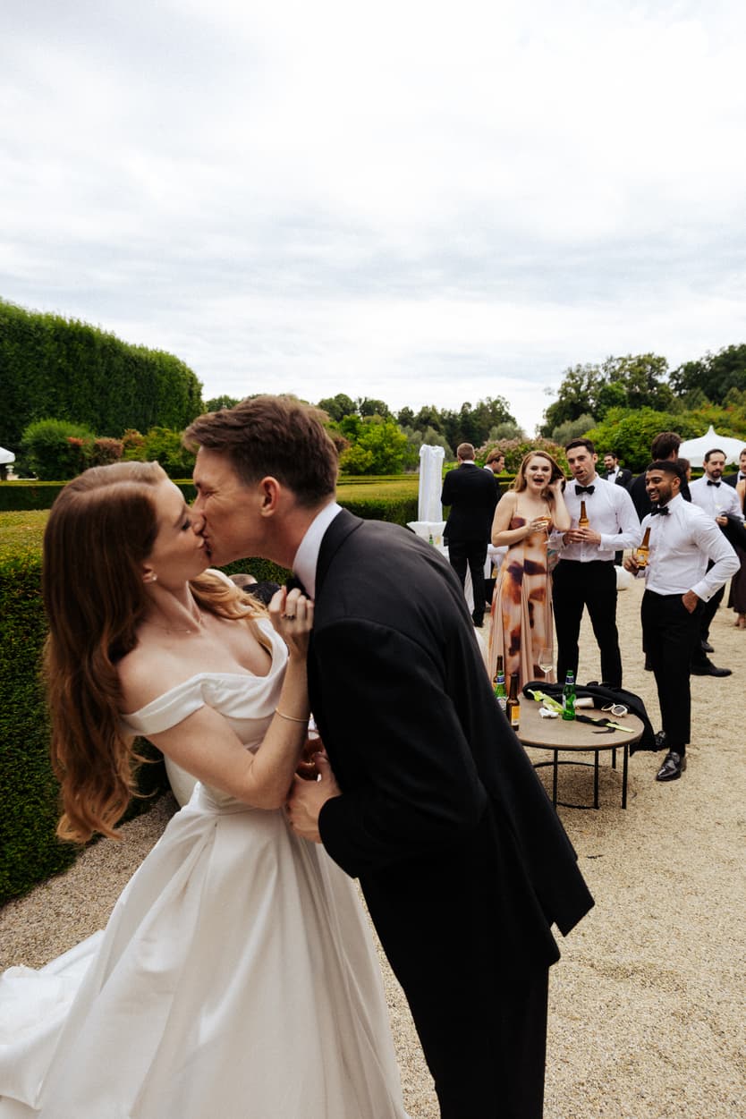 The couple kissing on the gravel path as their guests watch at the château cocktail hour
