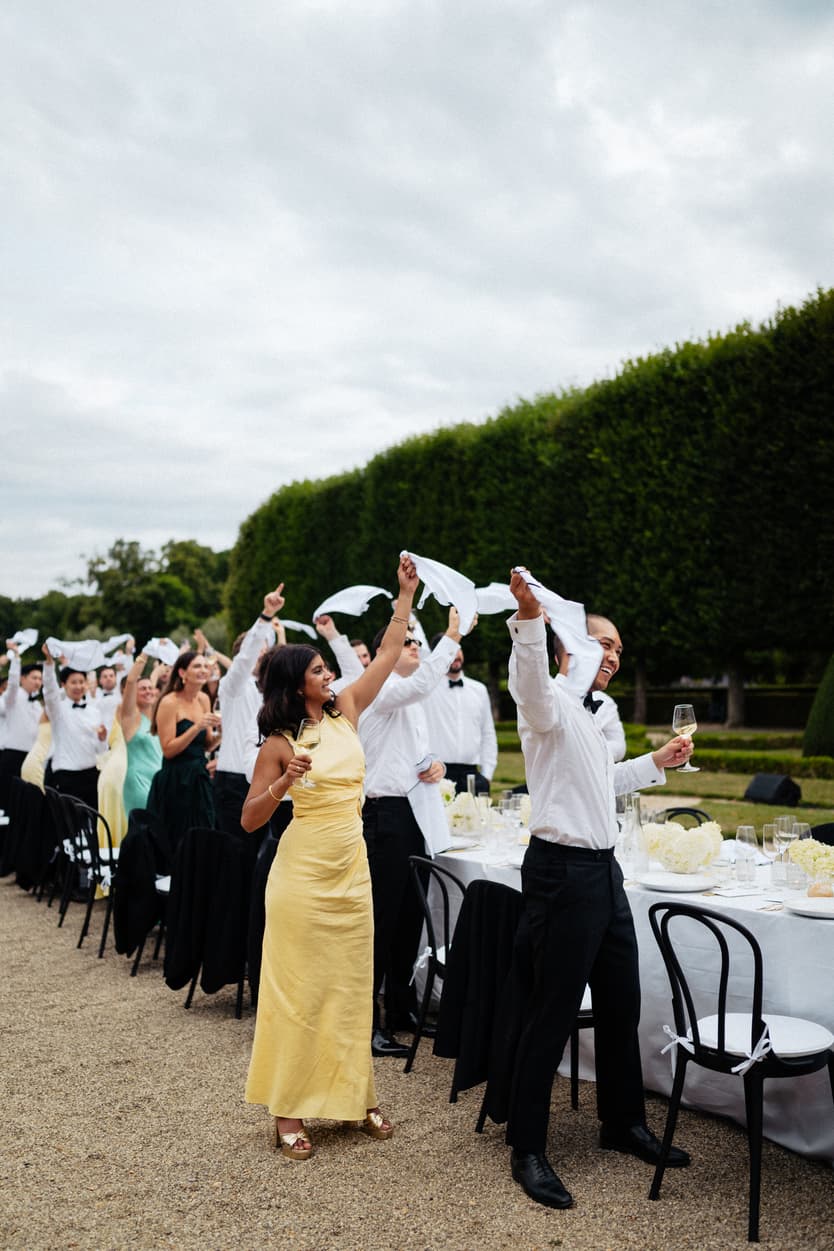 Guests on their feet waving napkins over the long wedding dinner table, white hydrangea down the middle