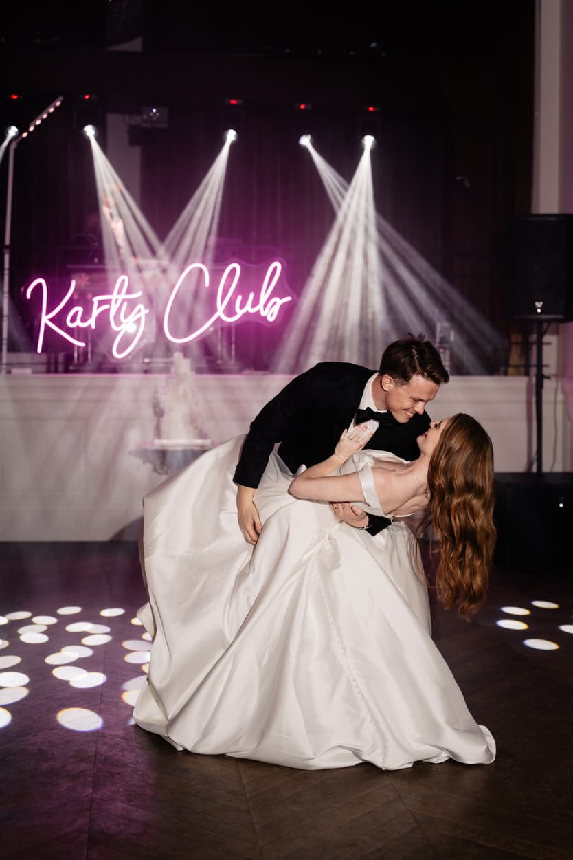 The couple in a dip on the lit dance floor, her satin skirt spread under the pink Karty Club neon sign