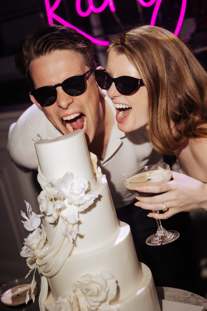 The couple in sunglasses over the tiered wedding cake and its sugar flowers, a cocktail in her hand, the neon sign above