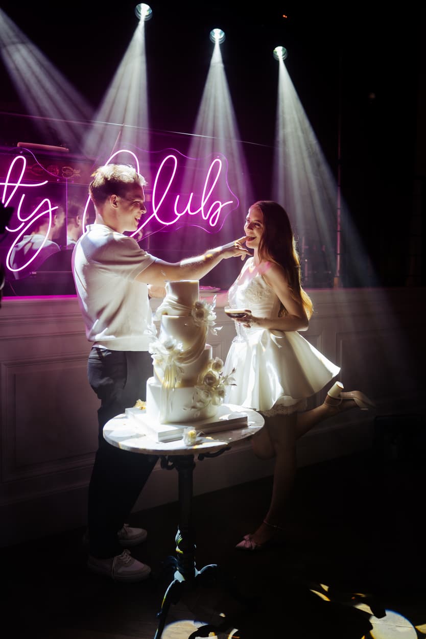 The groom feeding the bride a piece of the tiered wedding cake, her short satin second dress and the neon sign behind