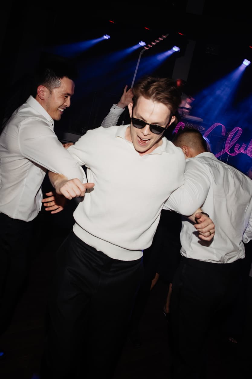 The groom in sunglasses pulled across the dance floor by his friends, blue beams and the neon sign behind