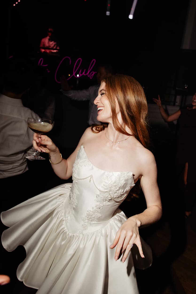 The bride laughing on the dance floor in her short satin dress, a coupe in her hand and the neon sign behind