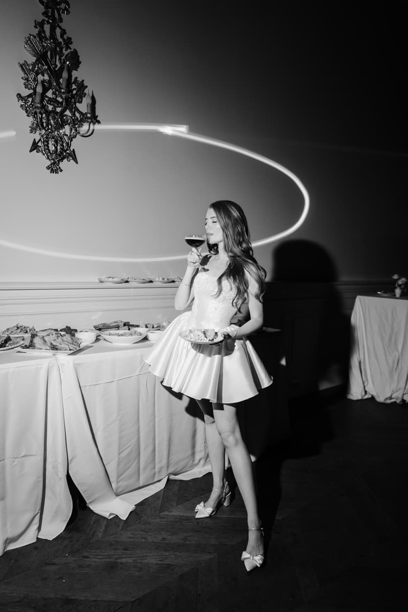 The bride in her short satin dress at the dessert table, a light trail curving under the chandelier, in black and white
