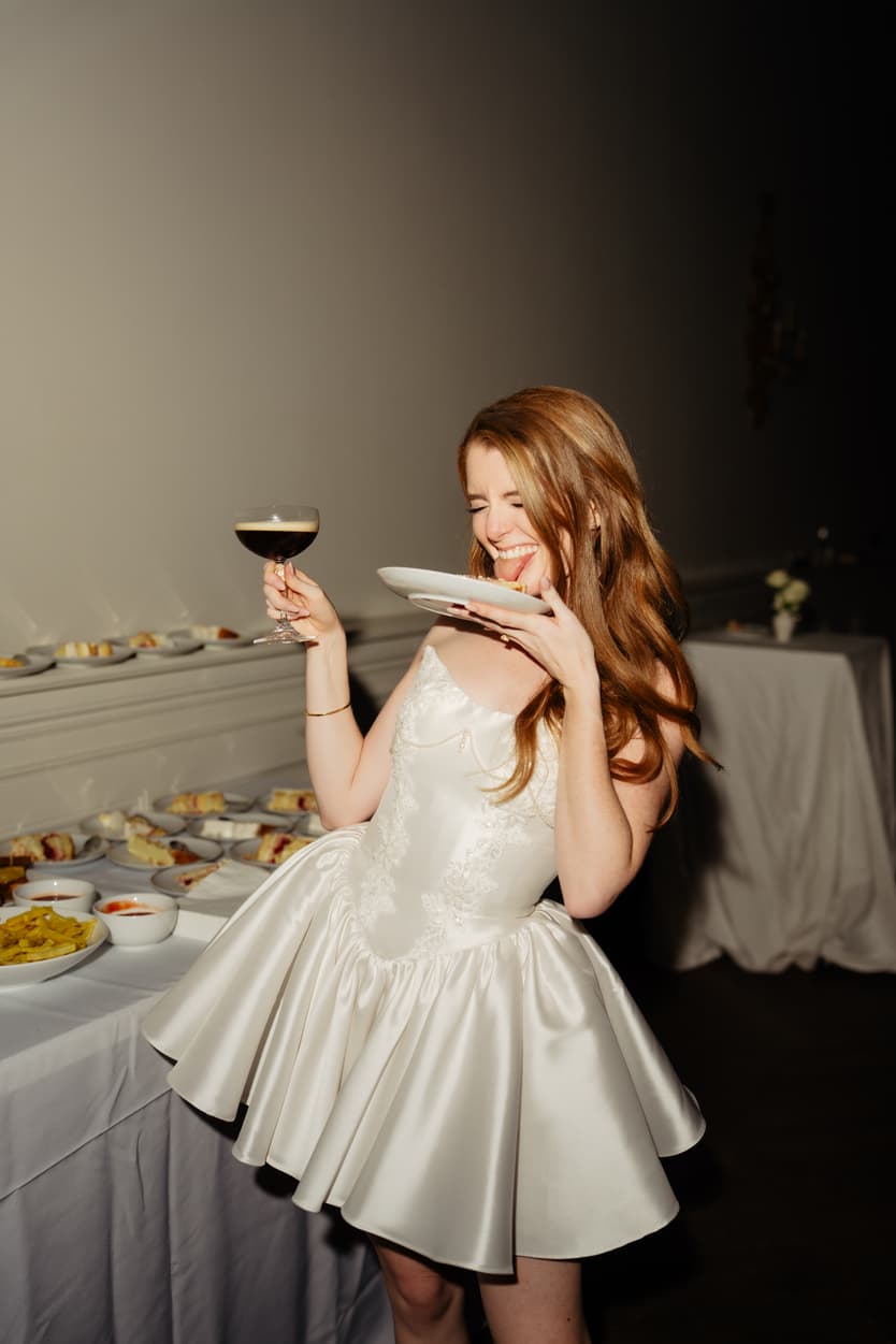 The bride in her short satin second dress laughing with a cocktail at the château’s dessert table