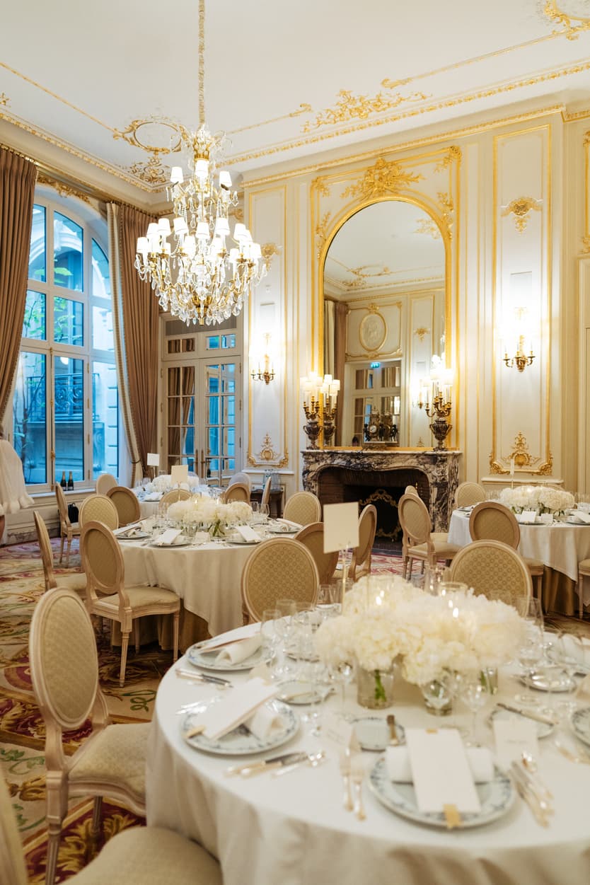 A wedding breakfast laid under the chandeliers of a gilded salon, white flowers on every round table
