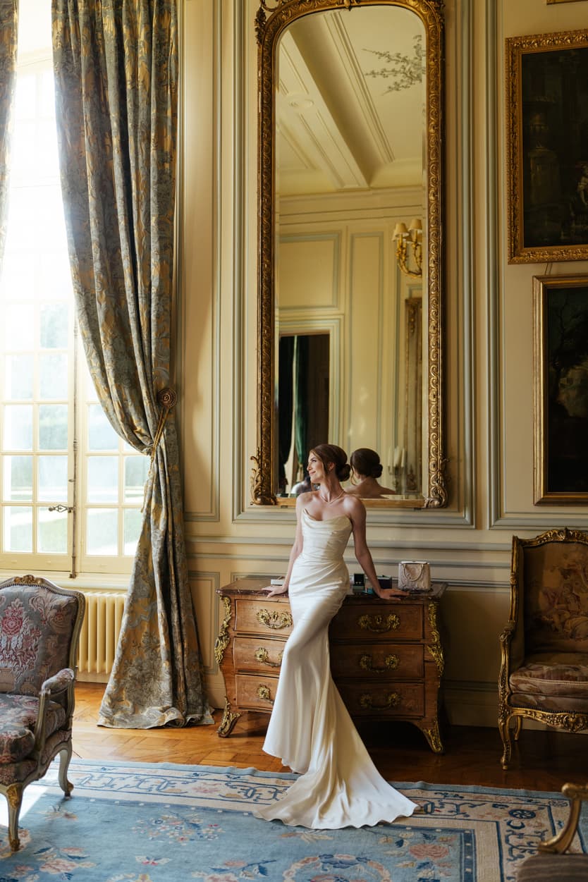 The bride in a white satin slip dress at the salon mirror of Château du Grand Lucé before the welcome dinner