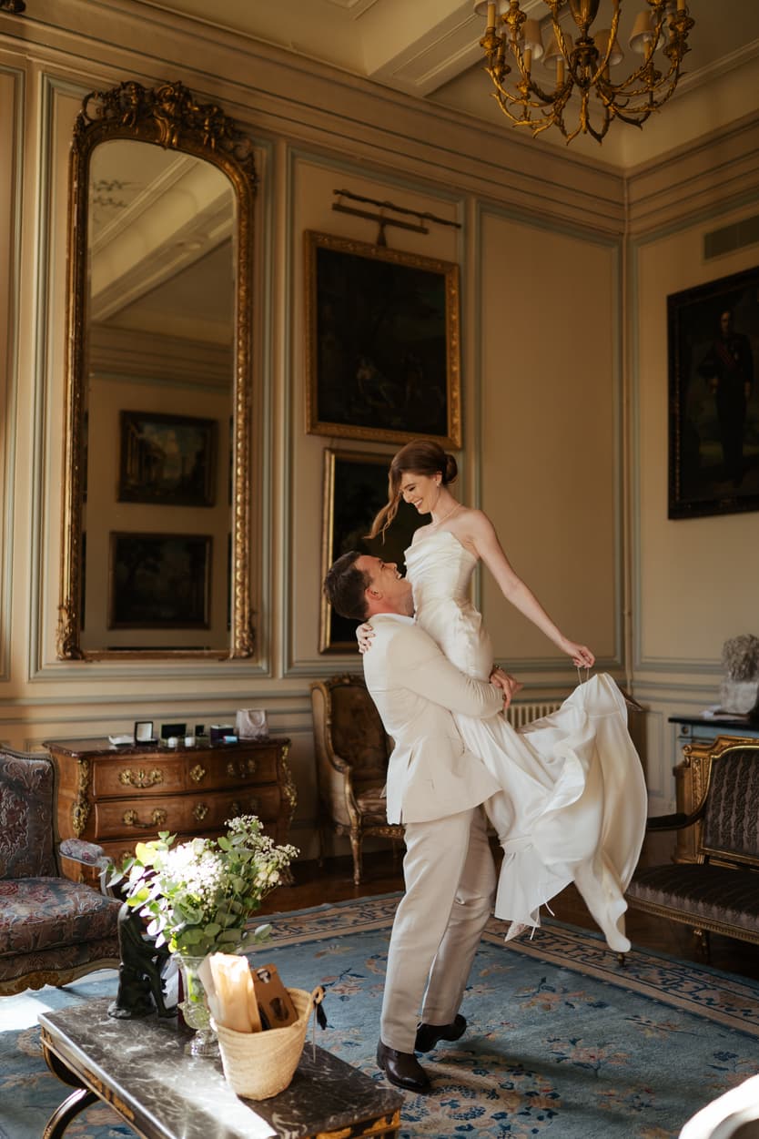 The groom lifting the bride in a château salon before the welcome dinner, her white satin slip dress swinging