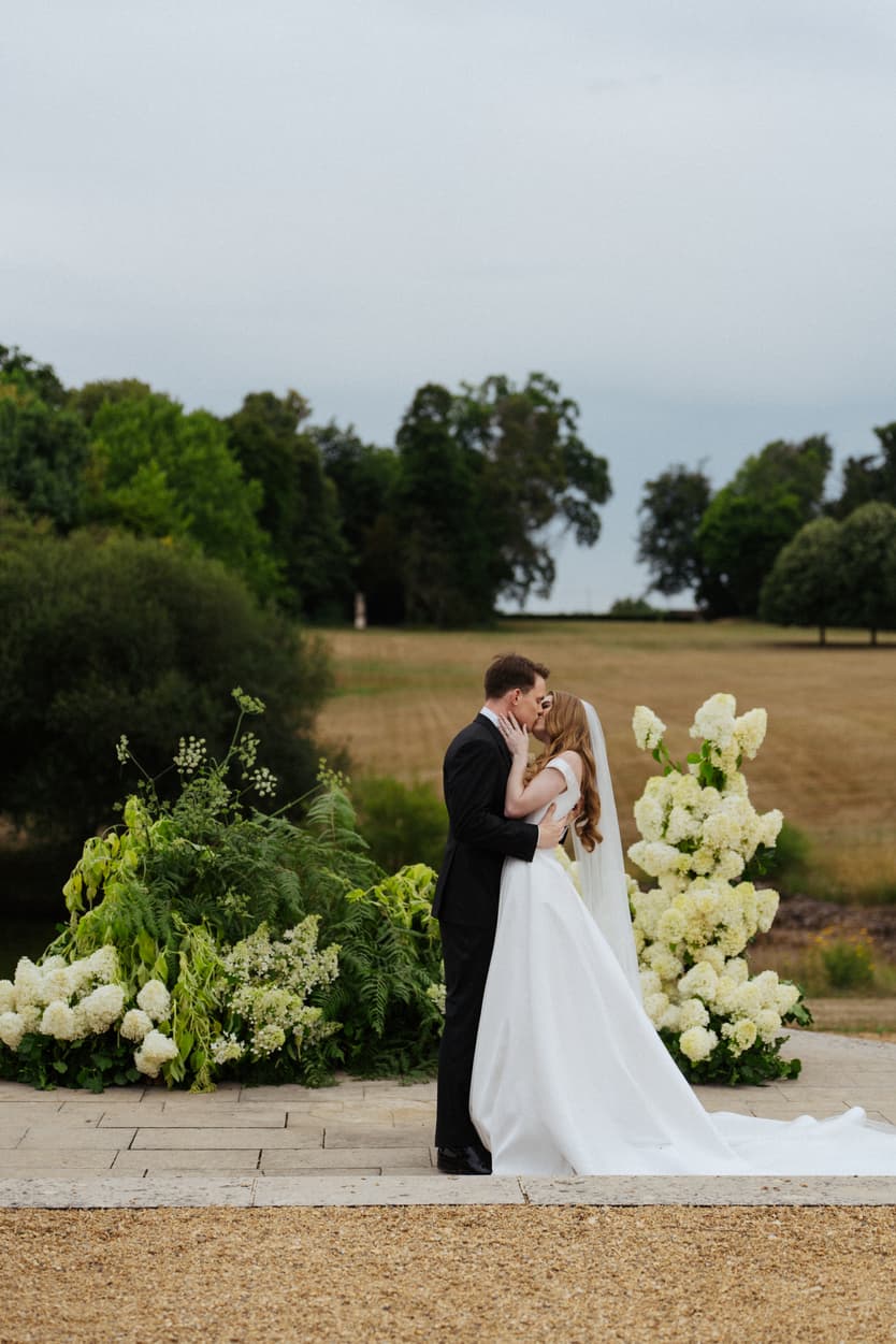 The couple’s first kiss beside the white hydrangea at the end of the aisle, her veil and satin train behind