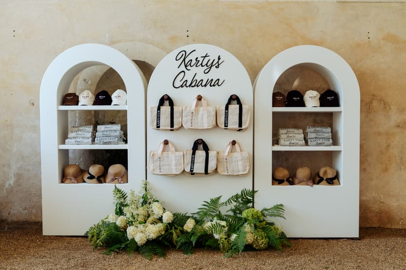 Karty’s Cabana at Château du Grand Lucé: caps, tote bags, towels and straw hats under three white arches, hydrangea below
