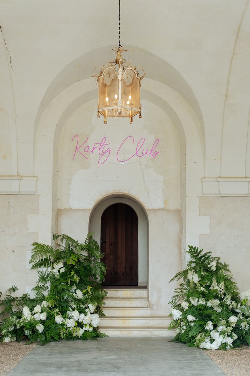The pink Karty Club neon sign over the orangerie door, a lantern above it and white hydrangea and ferns on the steps
