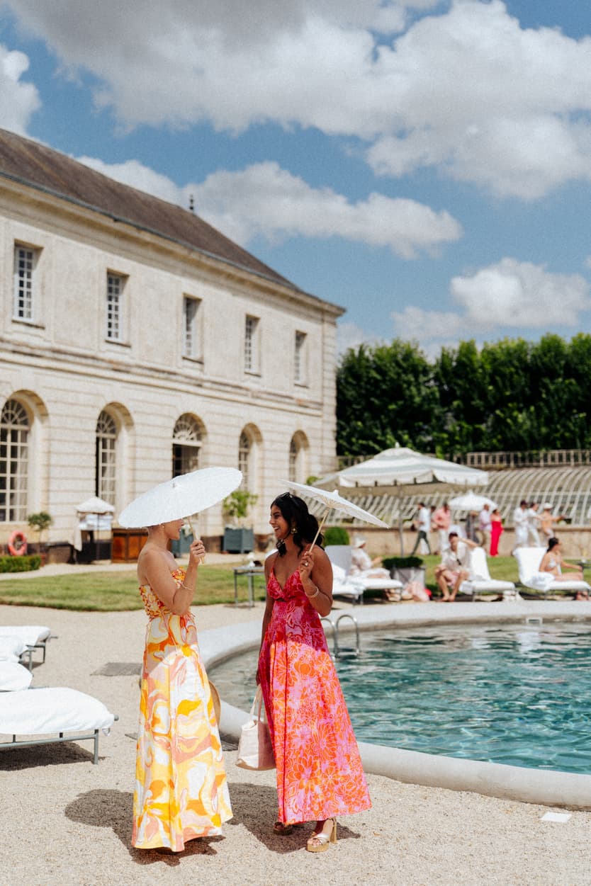 Two guests in bright summer dresses beside the pool at Château du Grand Lucé under a white parasol