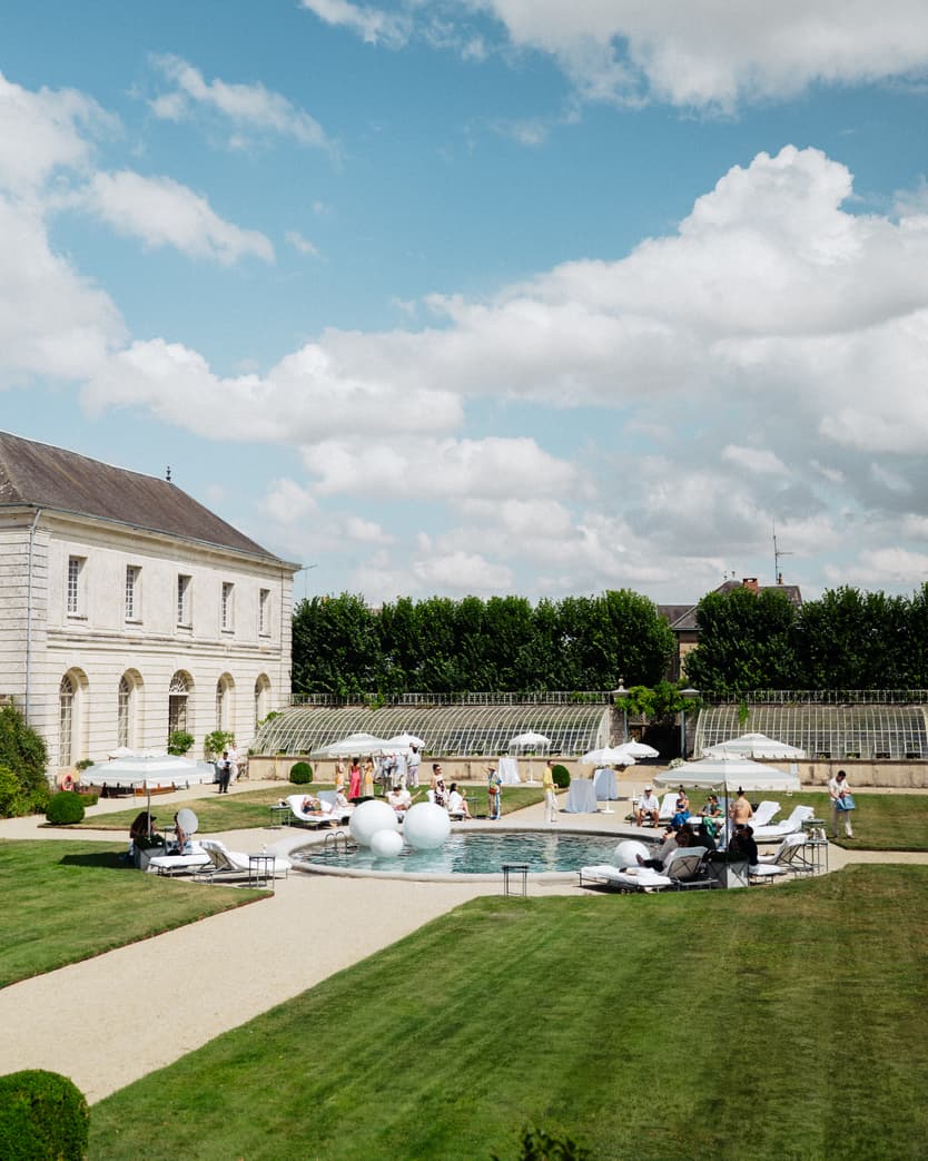 The pool day from the garden: loungers, white parasols and guests by the pool in front of the château’s orangerie